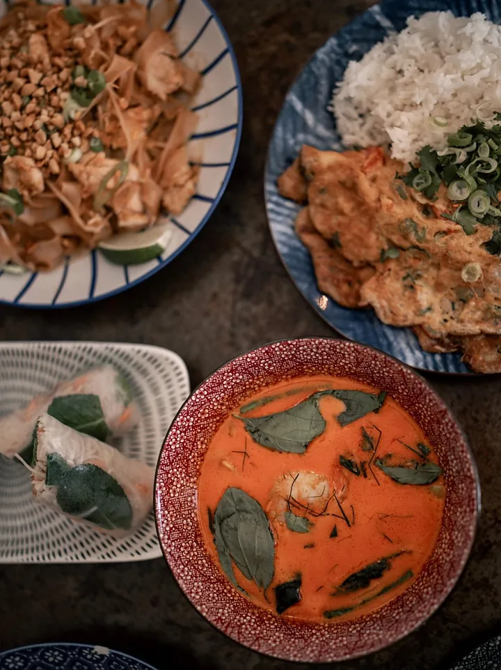 Vue du dessus de plusieurs plats : nouilles sautées aux cacahuètes, omelette avec riz, curry rouge au basilic et rouleaux de printemps.