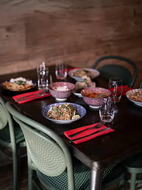 Une table sombre dressée avec des plats en bols, des serviettes rouges et des chaises vert sauge.