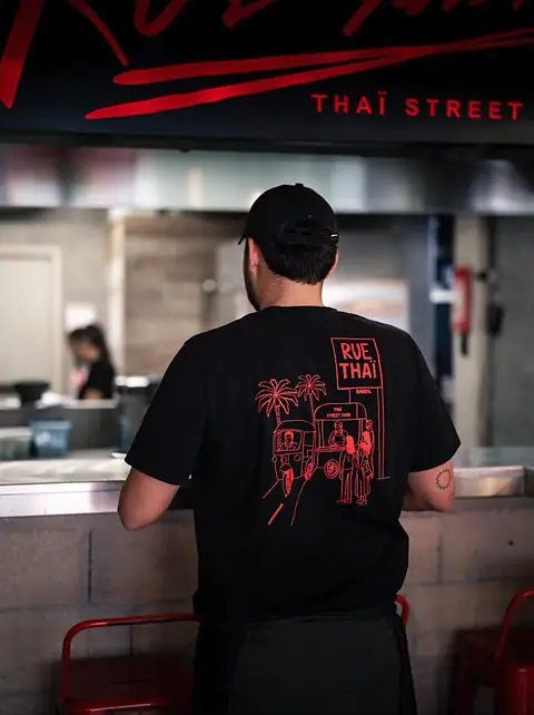 Un membre de l'équipe de dos, en casquette et T-shirt noir au logo, devant la cuisine ouverte.
